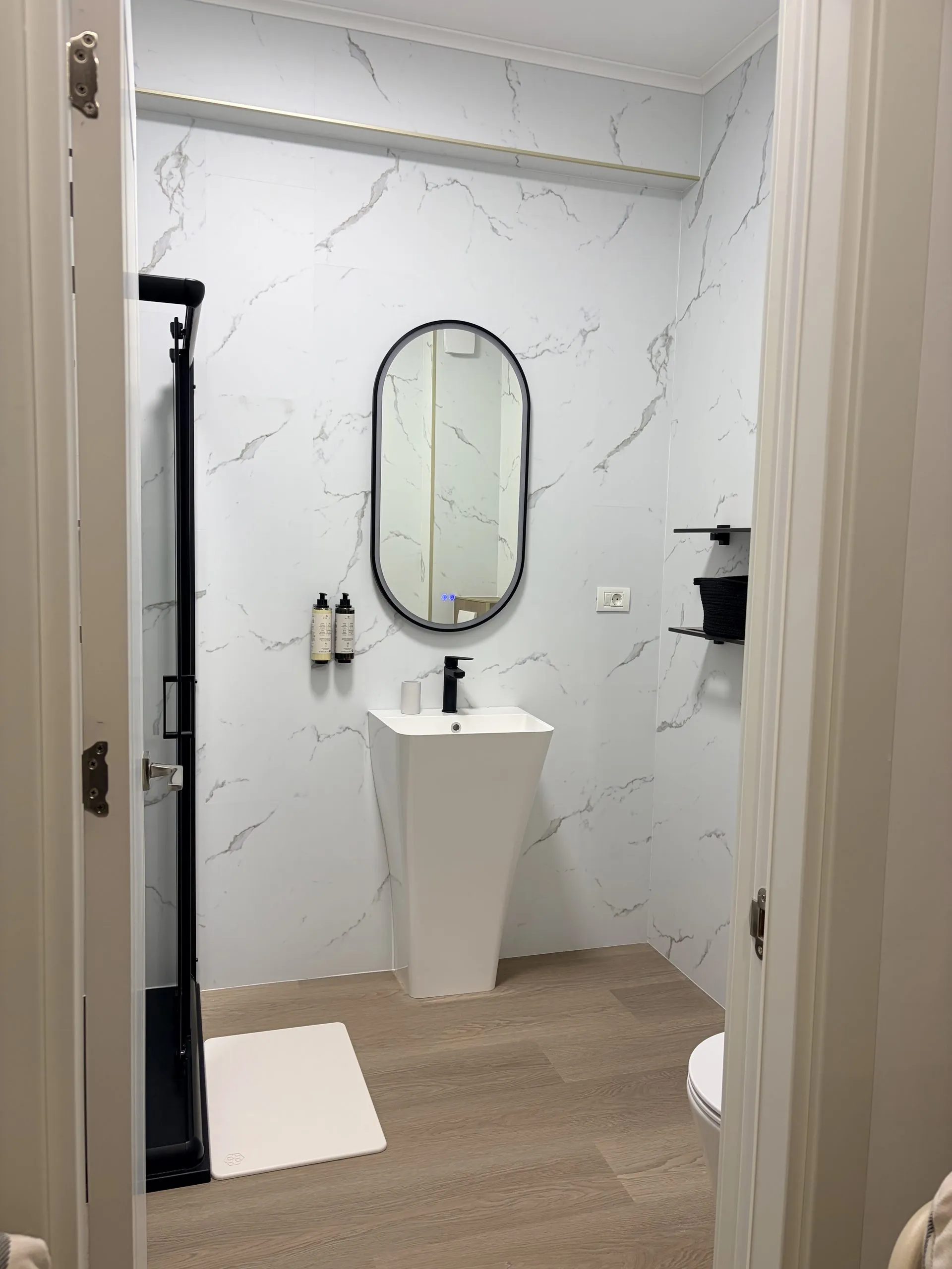 Bathroom seen through the open door — oval mirror, pedestal basin and walk-in shower beyond