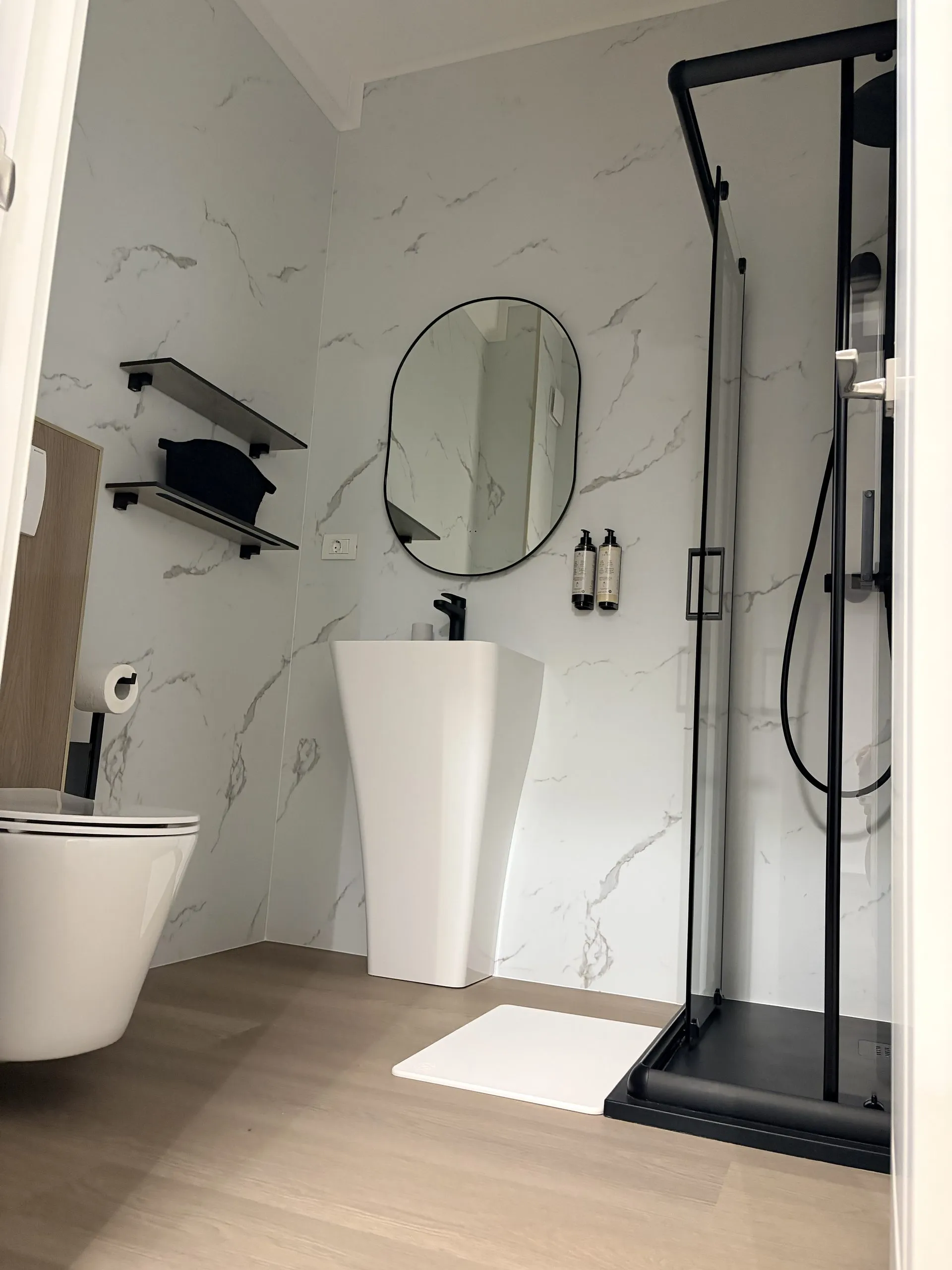 Bathroom lined in marble-look tile with an oval mirror, pedestal basin and a black-framed corner shower