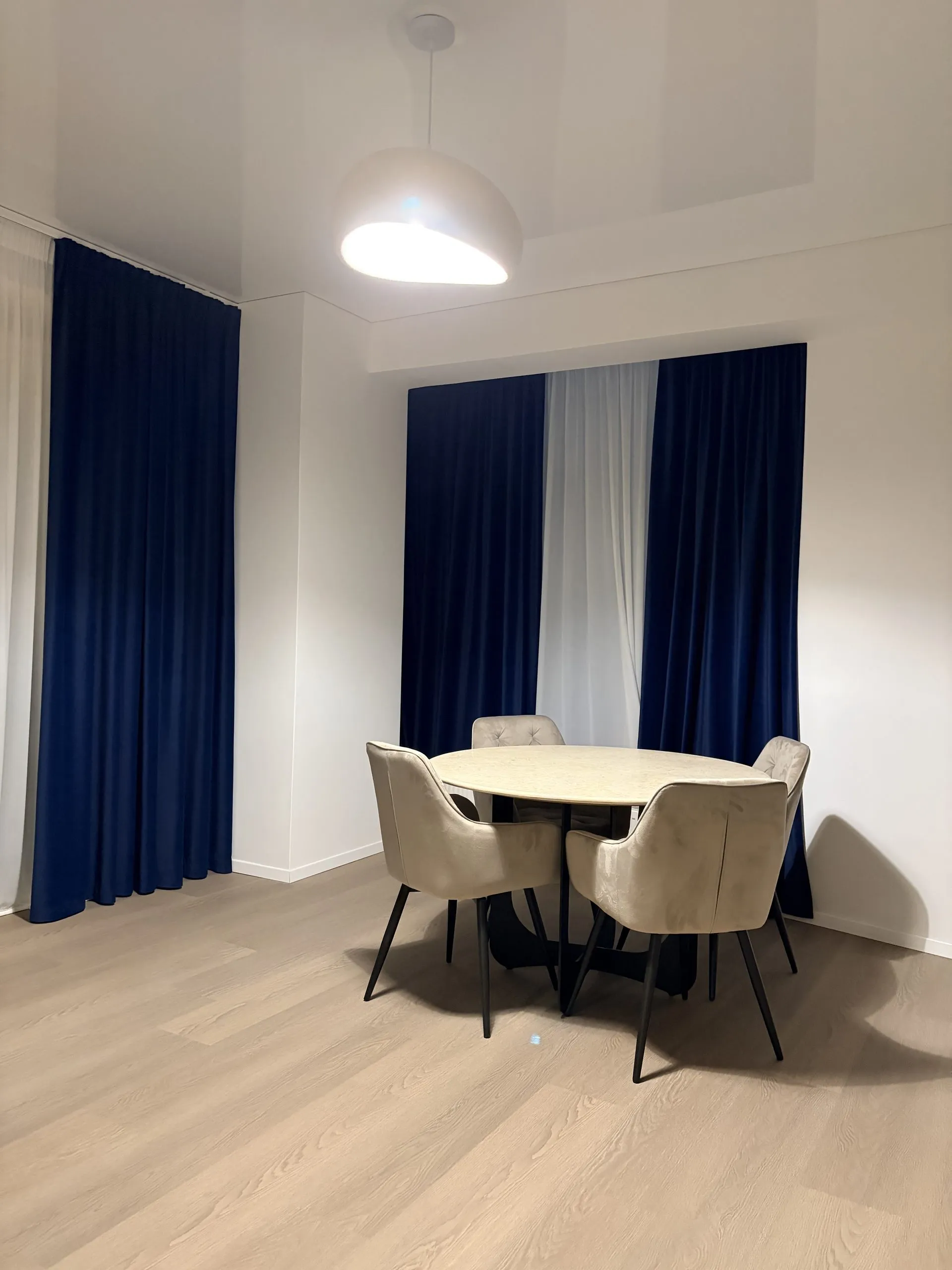Round dining table and four chairs beneath a pendant light, against navy and sheer curtains