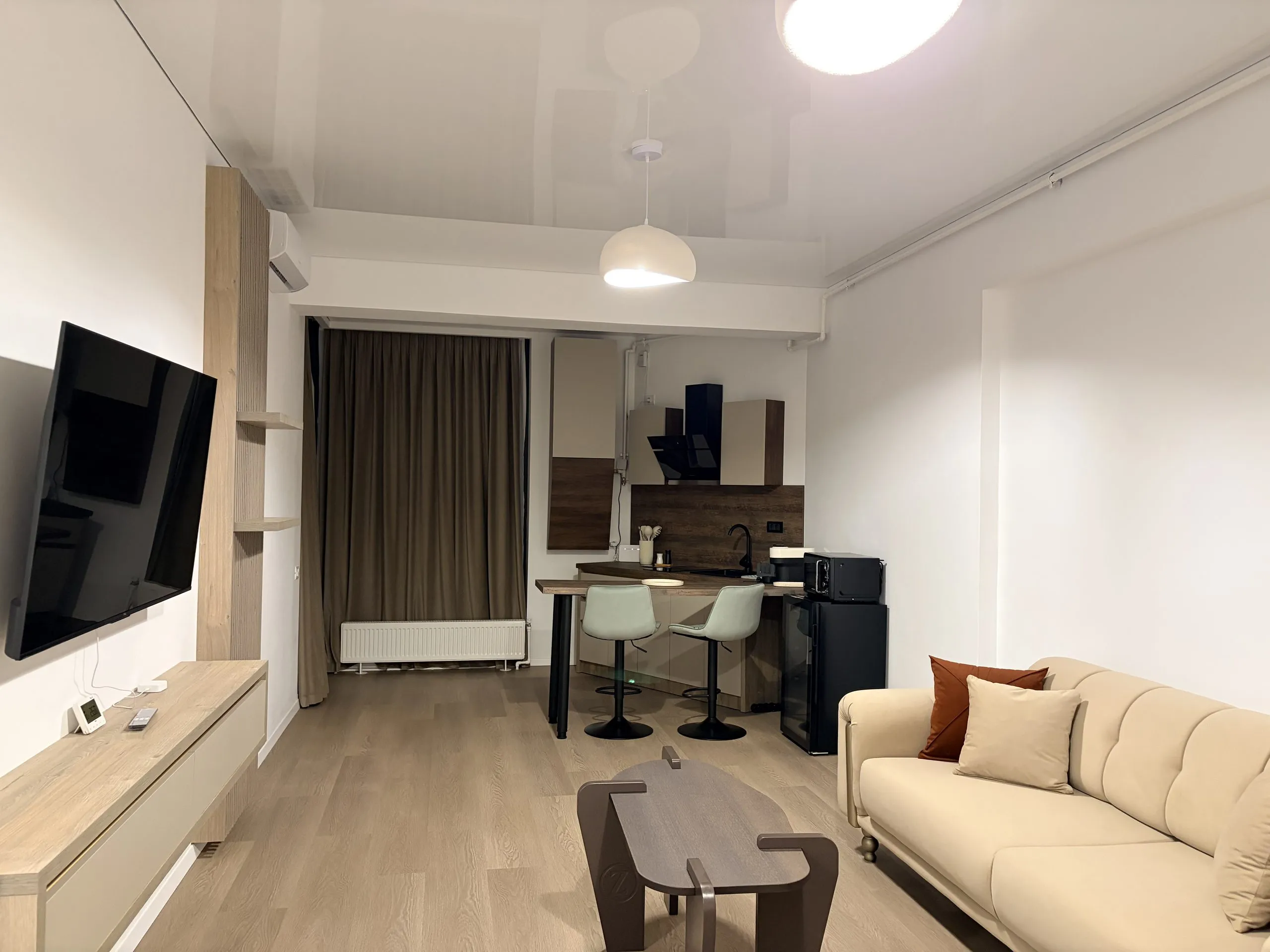 Open-plan apartment seen end to end — wall-mounted TV, kitchenette with a breakfast bar and stools, and a long cream sofa
